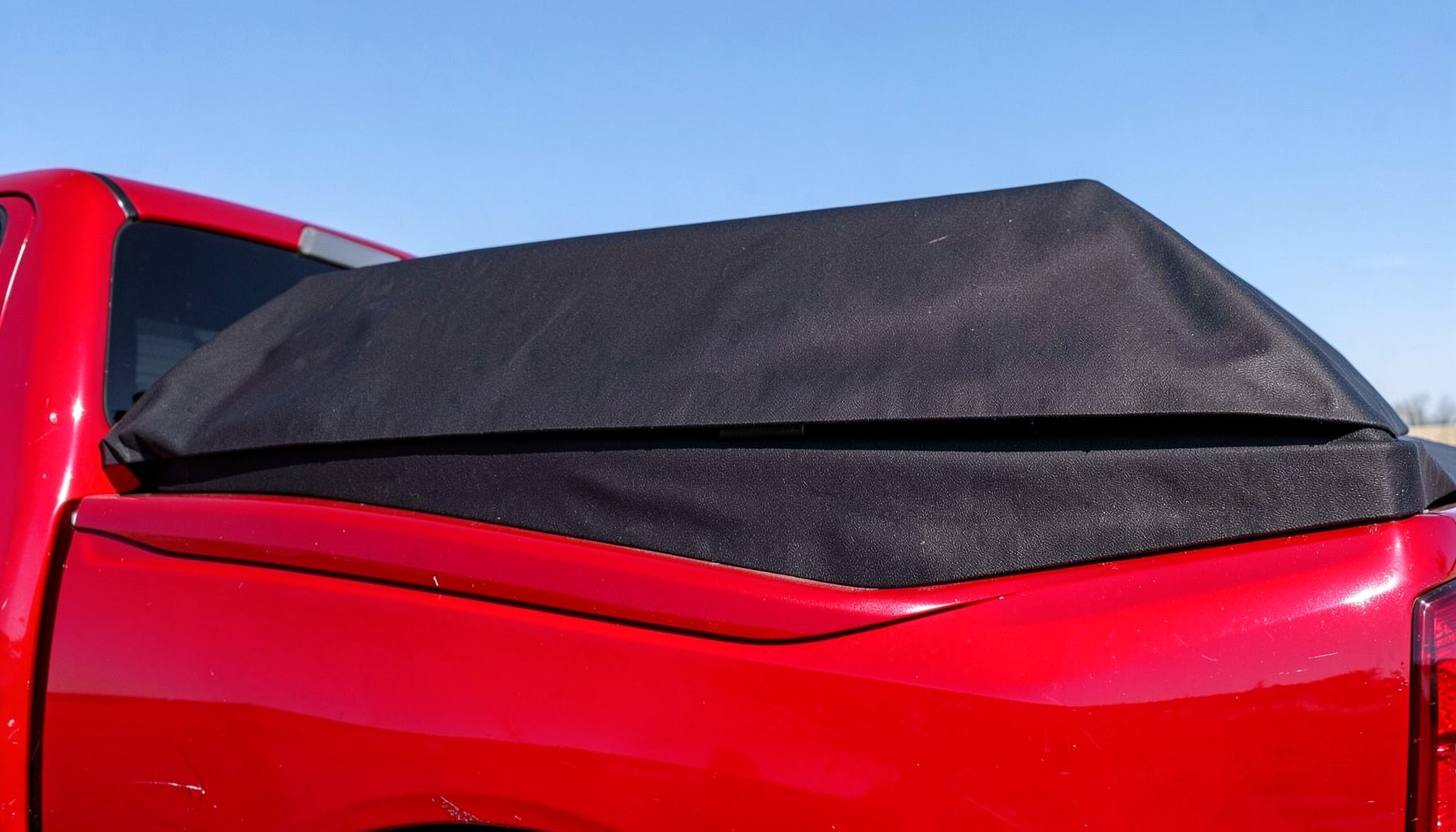 A folding hard tonneau cover in a tri-fold position on a red pickup truck, textured black panels under a clear sky