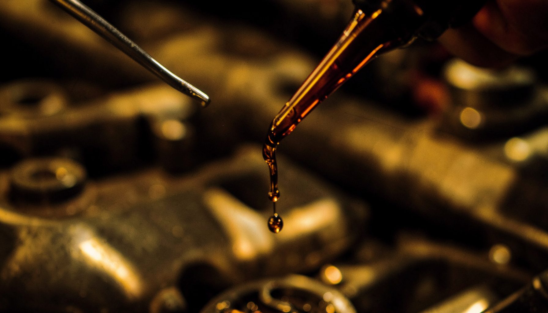 Close-up of dark amber motor oil dripping from a dipstick against a blurred metallic engine bay, warm golden-brown tones with deep shadows
