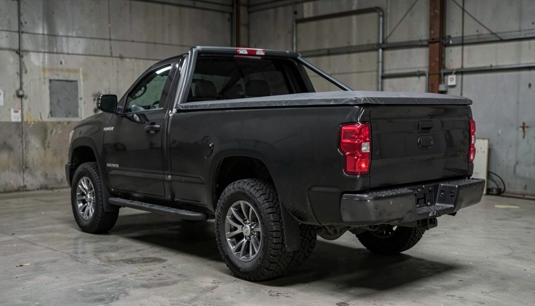 Low-profile roll-up tonneau cover installed on a dark pickup truck bed, seen from a rear three-quarter angle against a muted industrial backdrop