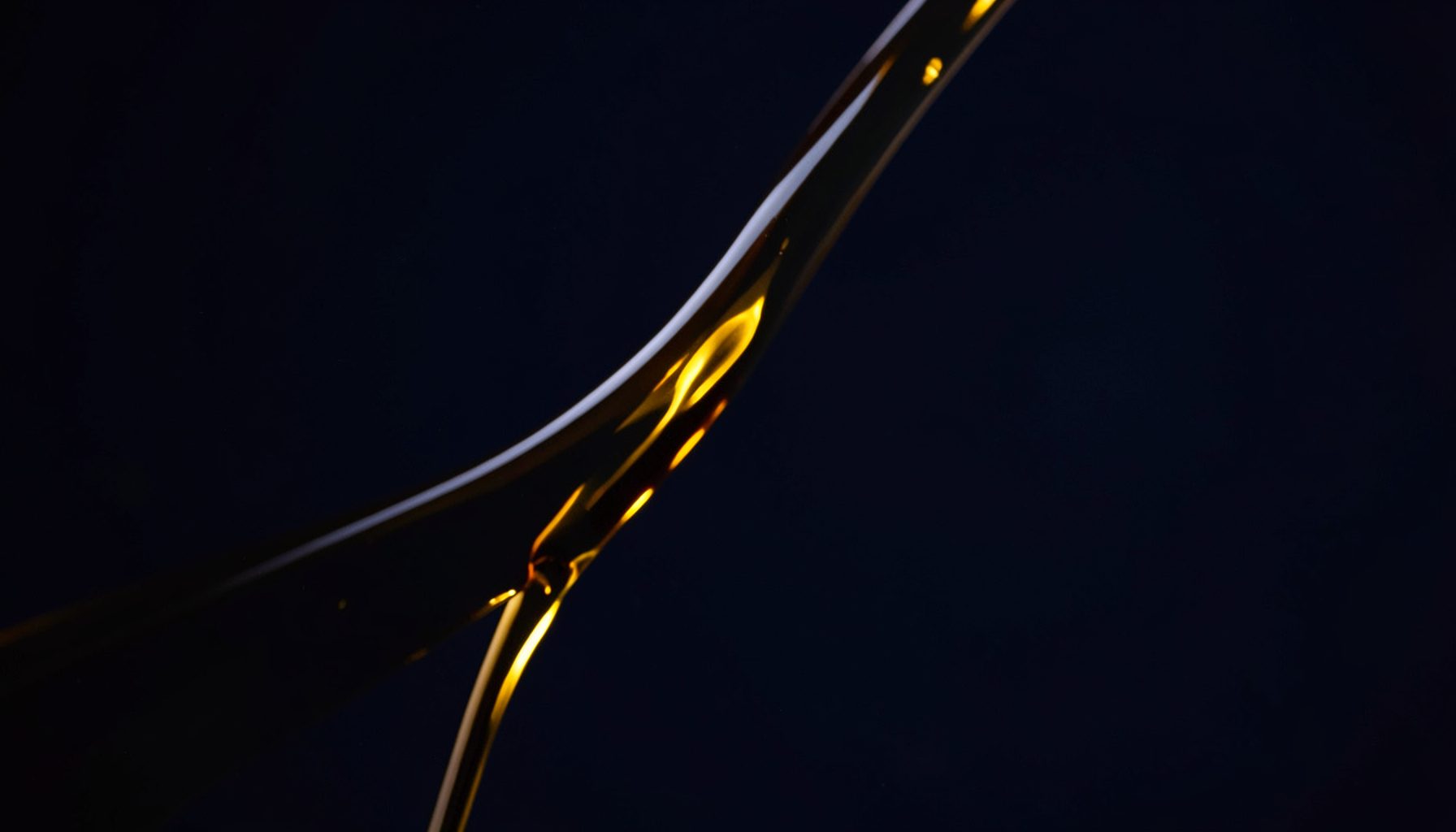 Warm amber-gold motor oil pouring in a smooth stream against a dark navy background, rich and glossy